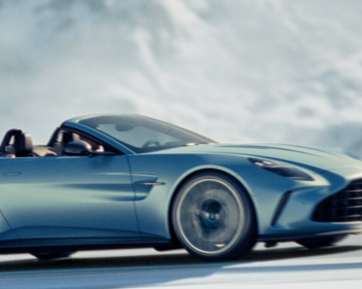 A blurred action shot of a blue convertible Aston Martin speeding across a snowy or icy landscape with a cloudy sky.