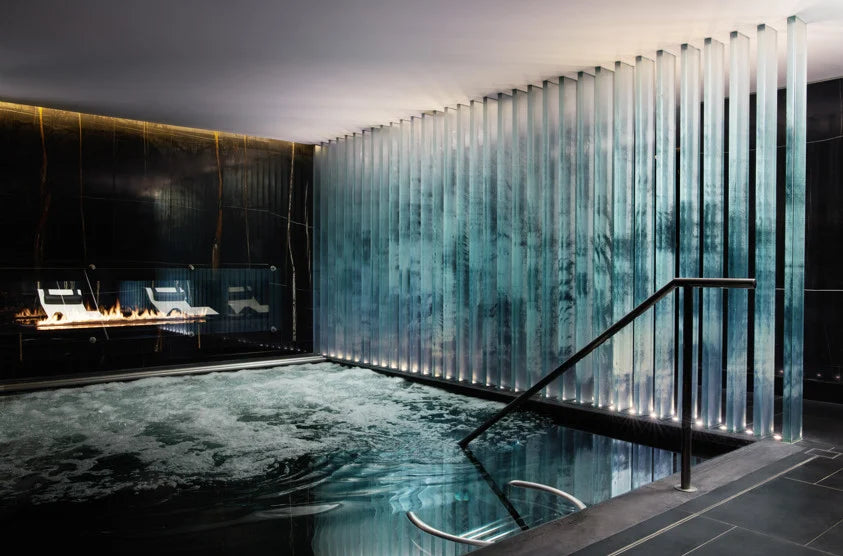 A modern indoor swimming pool surrounded by dark walls and a wall of vertical glass panels, creating a waterfall-like effect.