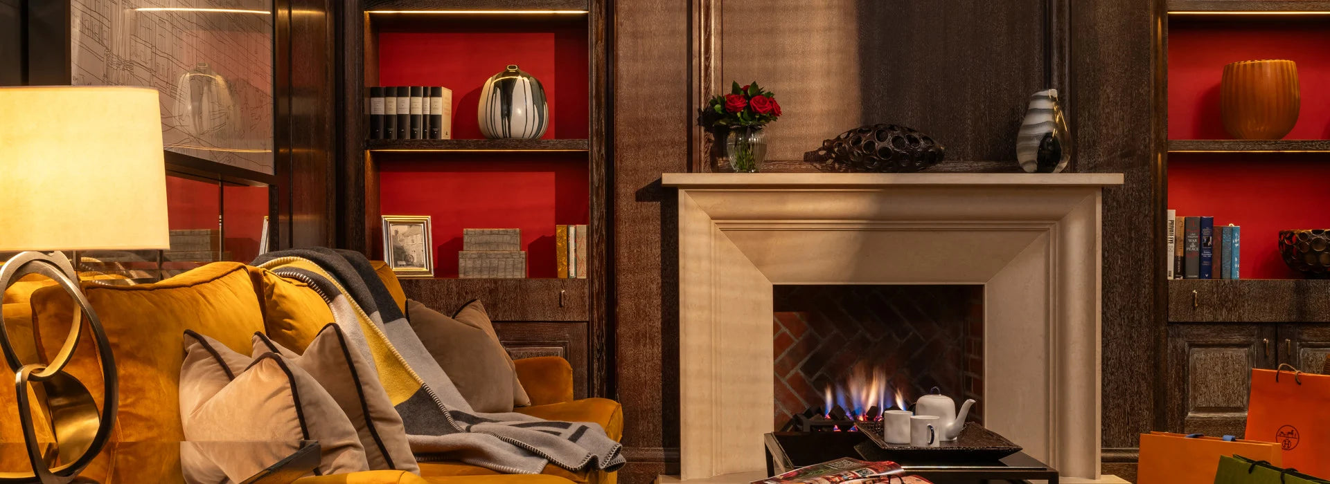 A cosy living room with a lit fireplace, yellow sofa with throw, pillows and a blanket, wooden walls, and built-in bookshelves with red back panels.