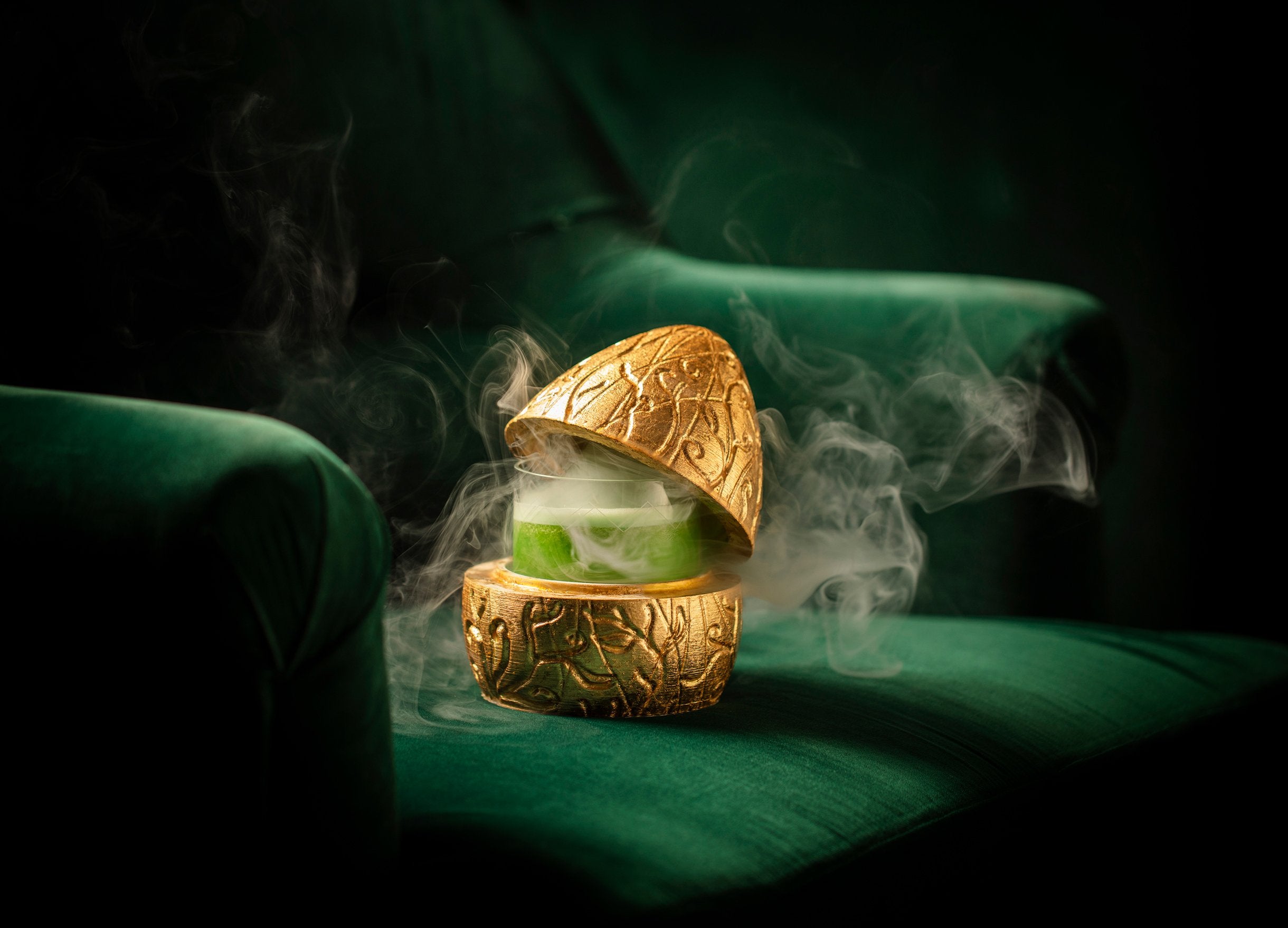 A smoking cocktail served in an ornate gold eggshell-shaped glass, resting on a deep green velvet armchair, at The Donovan Bar in London.