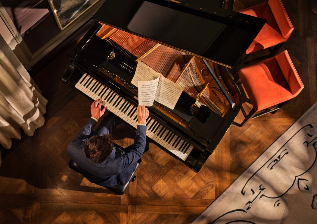 An aerial view of a person in a blue suit playing a grand piano on a herringbone wood floor.