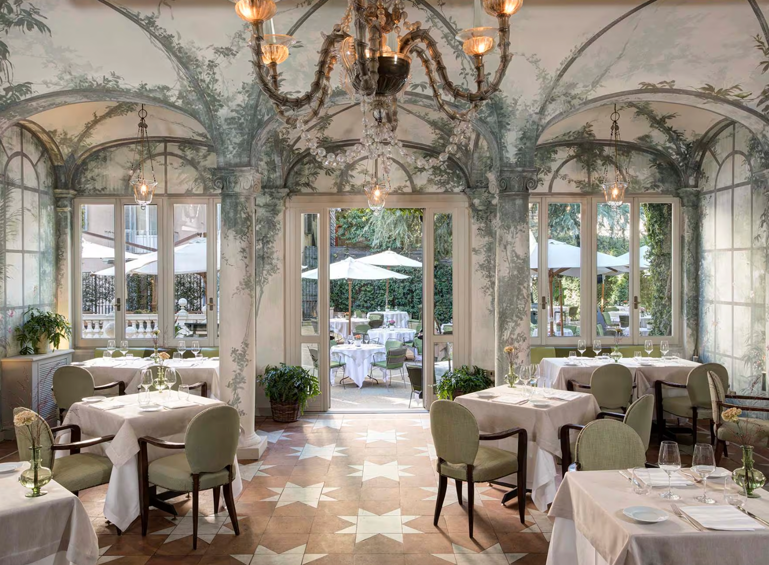 A beautifully decorated dining room with a hand-painted mural of intertwined trees and vines on the walls and arched ceiling, leading out to an outdoor patio with white tables and umbrellas.