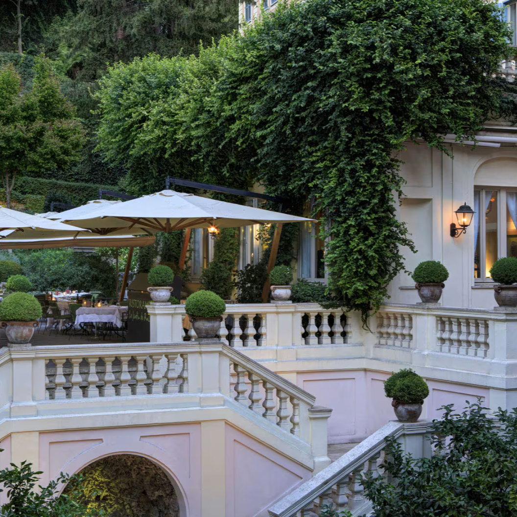 A luxurious hotel terrace with a grand staircase, lush green ivy and trees, and dining tables set up under large umbrellas.
