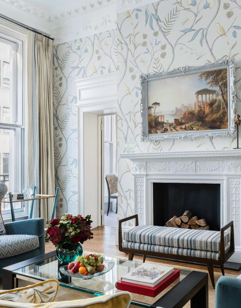 A bright, traditional living room within a suite at Brown's Hotel with floral wallpaper, a white marble fireplace and elegant furniture.