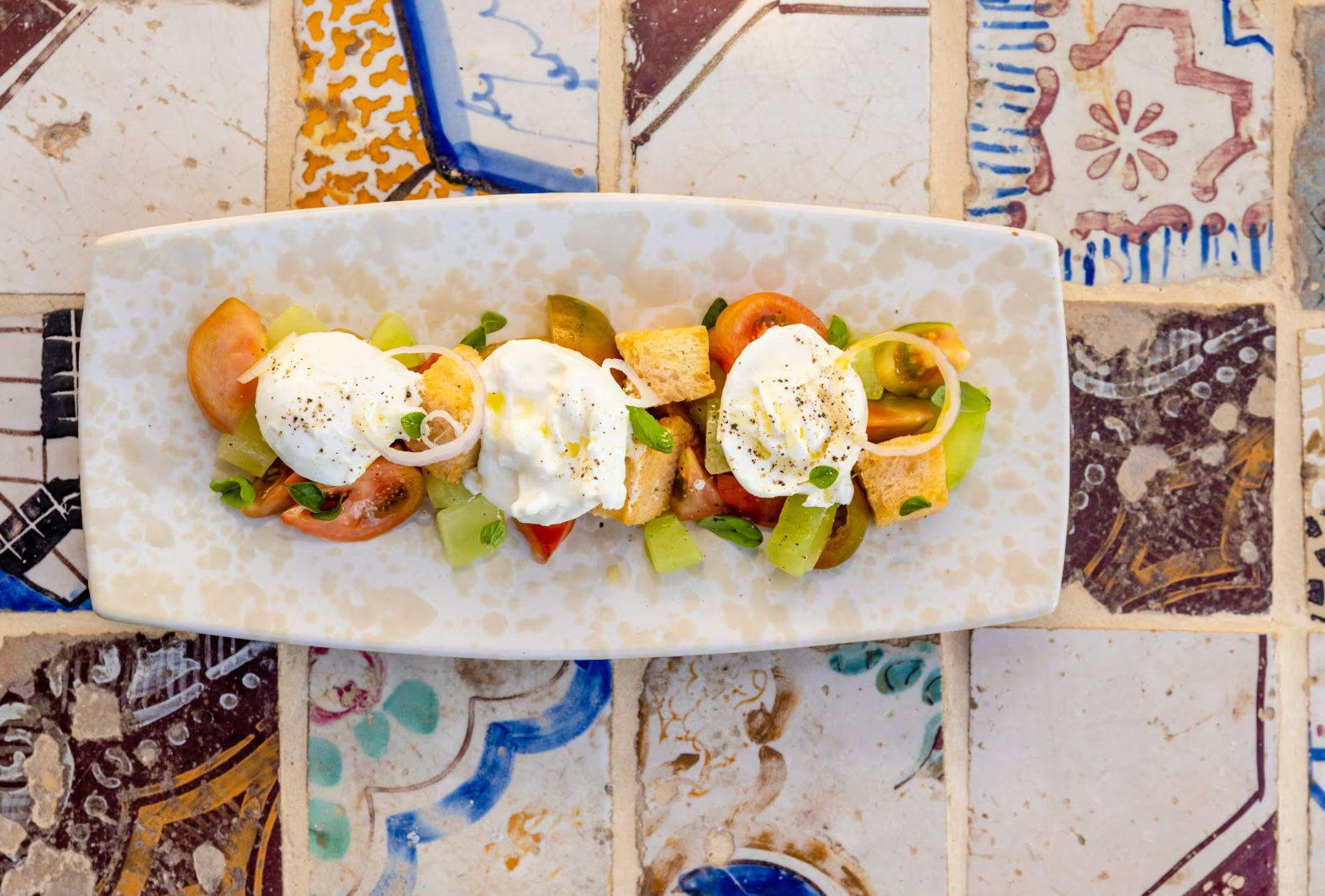 A vibrant salad with burrata, tomatoes, and cucumbers on a mosaic-tiled table.