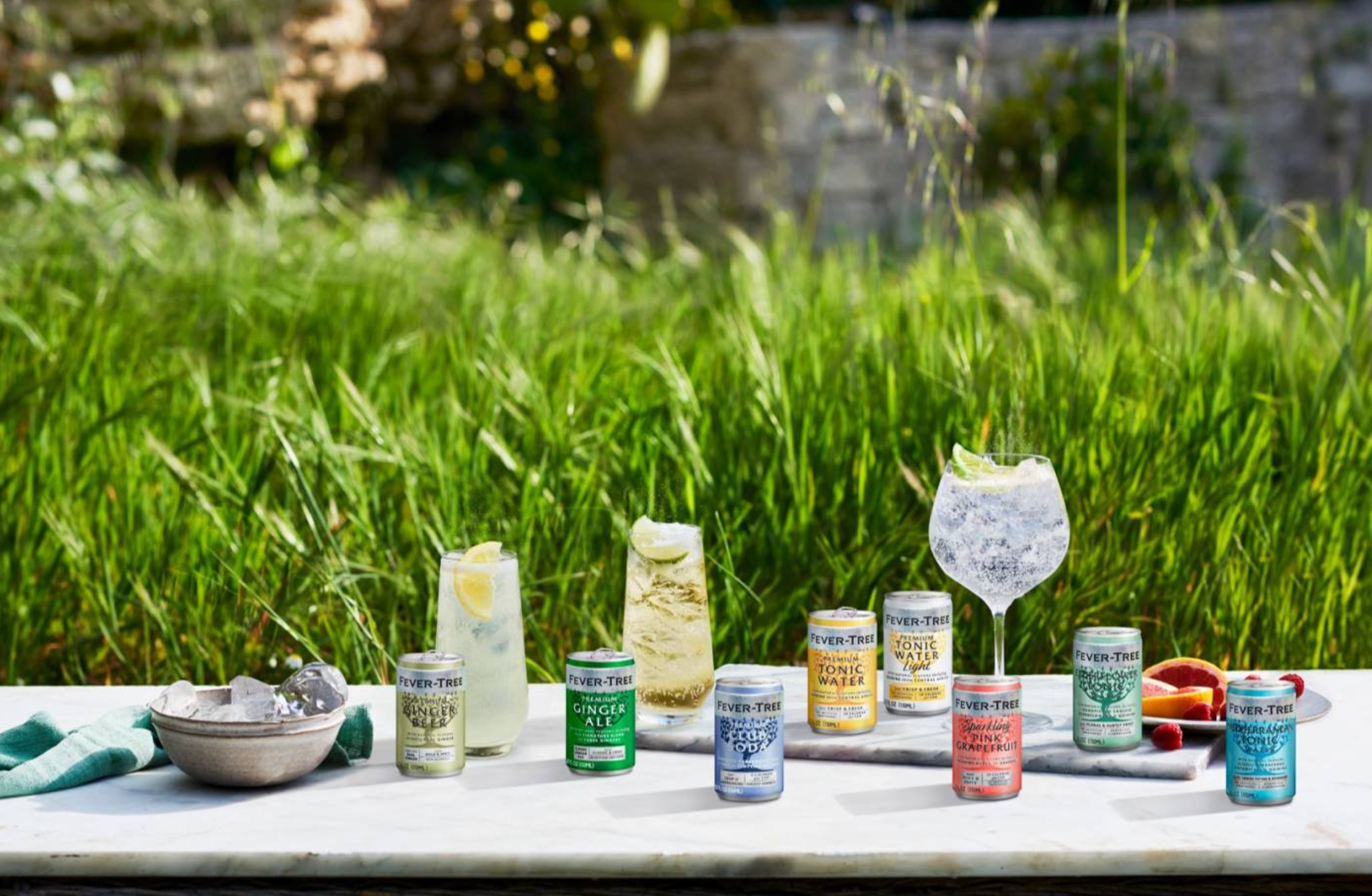 A selection of Fever-Tree beverages in cans and glasses on a marble table outdoors, surrounded by tall green grass.