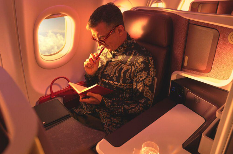 A man wearing glasses is seated in a window seat on a plane, writing in a small notebook during golden hour.