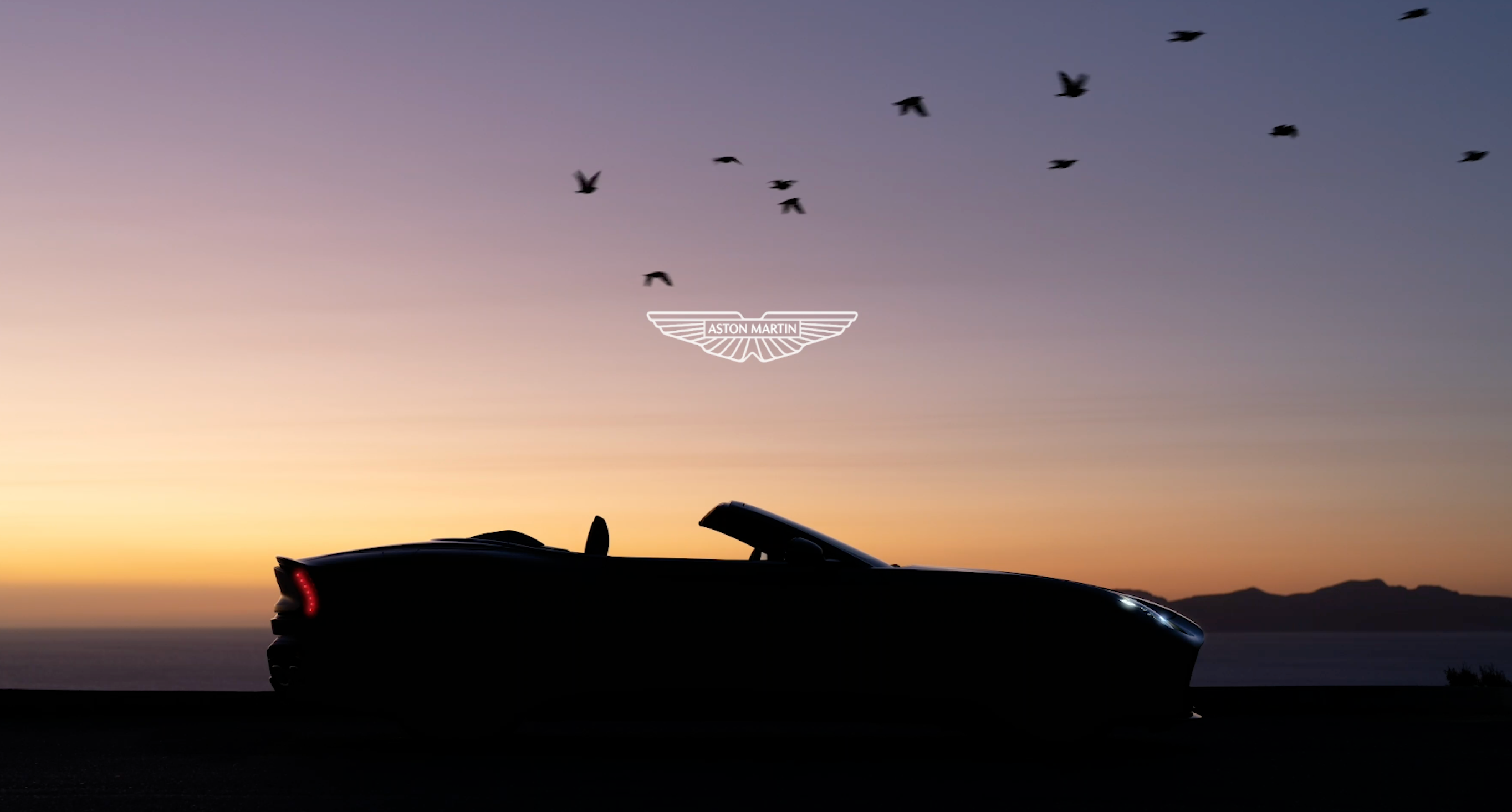 A silhouette of a convertible Aston Martin sports car at sunset, with birds flying overhead and the brand's logo in the sky.