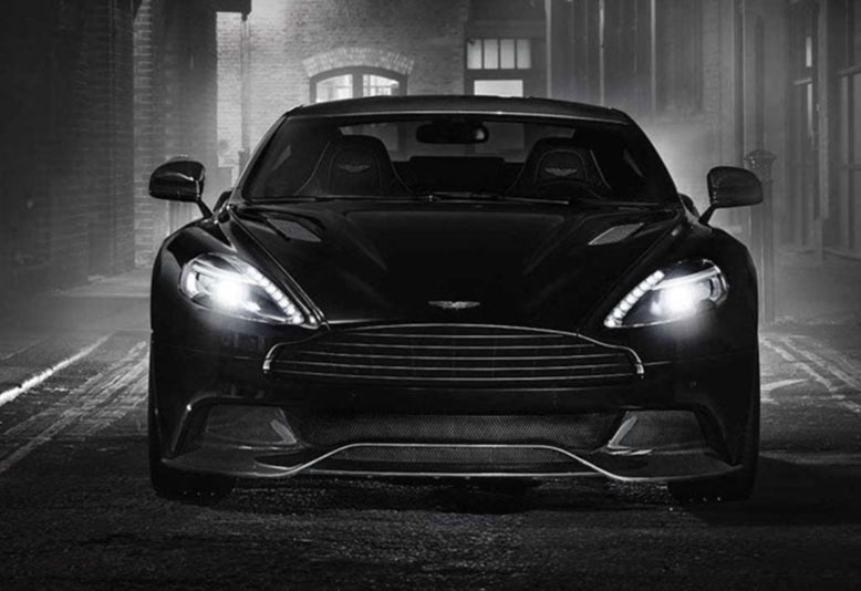 A striking, front-facing view of a dark Aston Martin sports car with its headlights on, parked on a cobblestone street at night.
