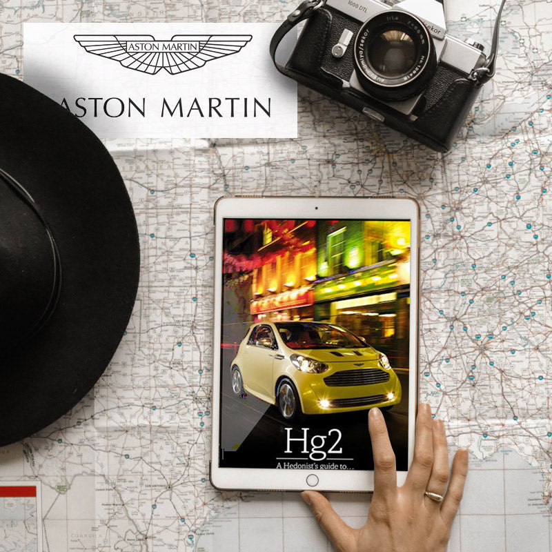 A person's hand is holding a tablet that displays an image of a yellow Aston Martin car driving at night. The tablet, along with a vintage camera, a black fedora, and an Aston Martin paper sign, is arranged on top of a map.