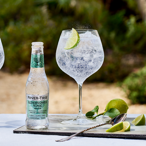 A balloon-shaped glass, filled with gin and tonic, ice, and a slice of lime next to a bottle of Fever-Tree Elderflower Tonic Water